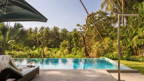 A swimming pool with a lounge chair and umbrella, surrounded by lush greenery. - Viraam Estate by RÊVE/Infinity heated pool with caretaker/Chef (Assagao)