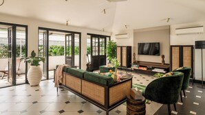 A spacious living room with a large sofa, a television, and a coffee table. There are several chairs, a potted plant, and a guitar. The room has large windows and an air conditioning unit on the wall. - Viraam Estate by RÊVE/Infinity heated pool with caretaker/Chef (Assagao)