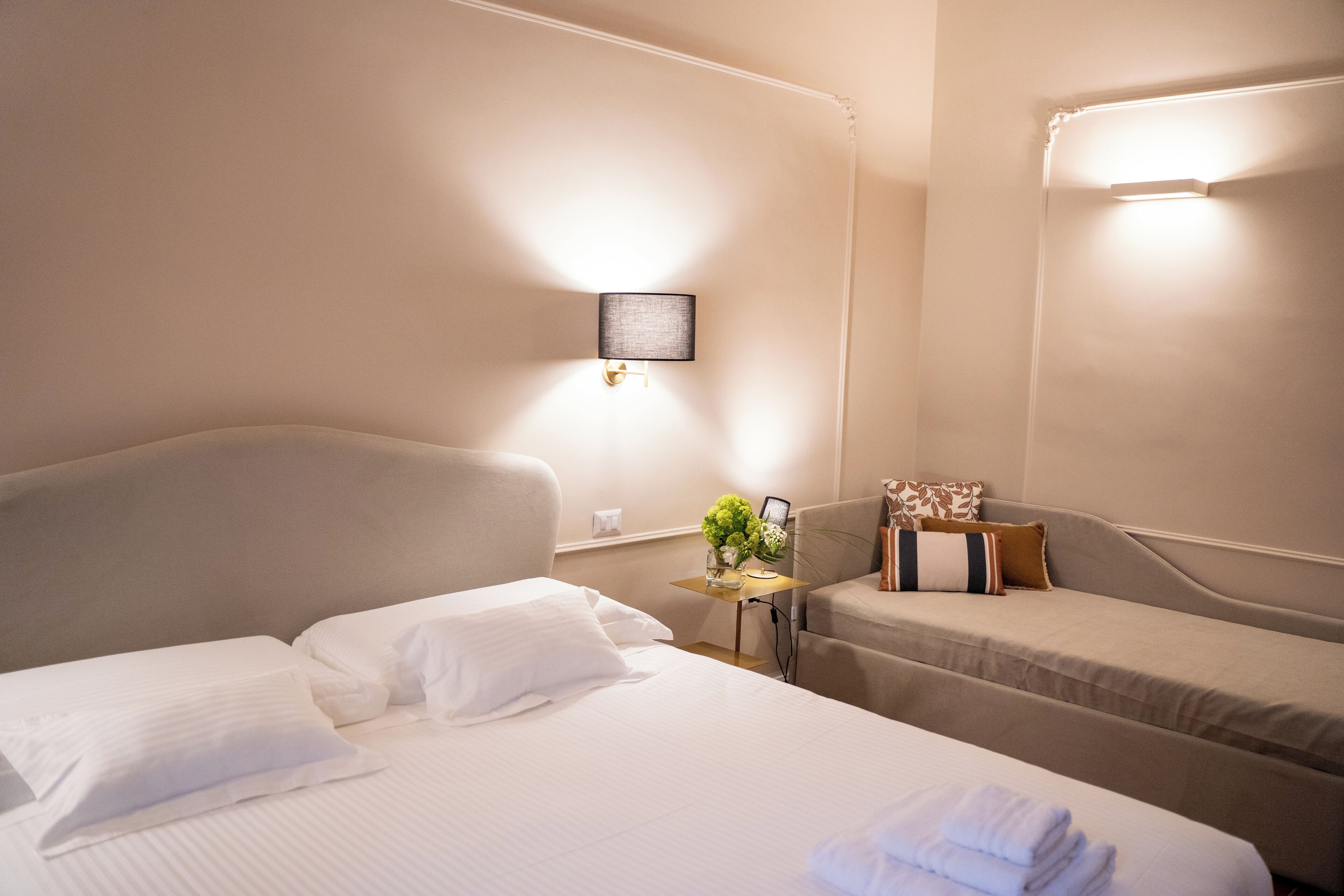 Habitación de hotel con una cama grande, una cama individual, una mesita de noche con una lámpara e iluminación empotrada en la pared.