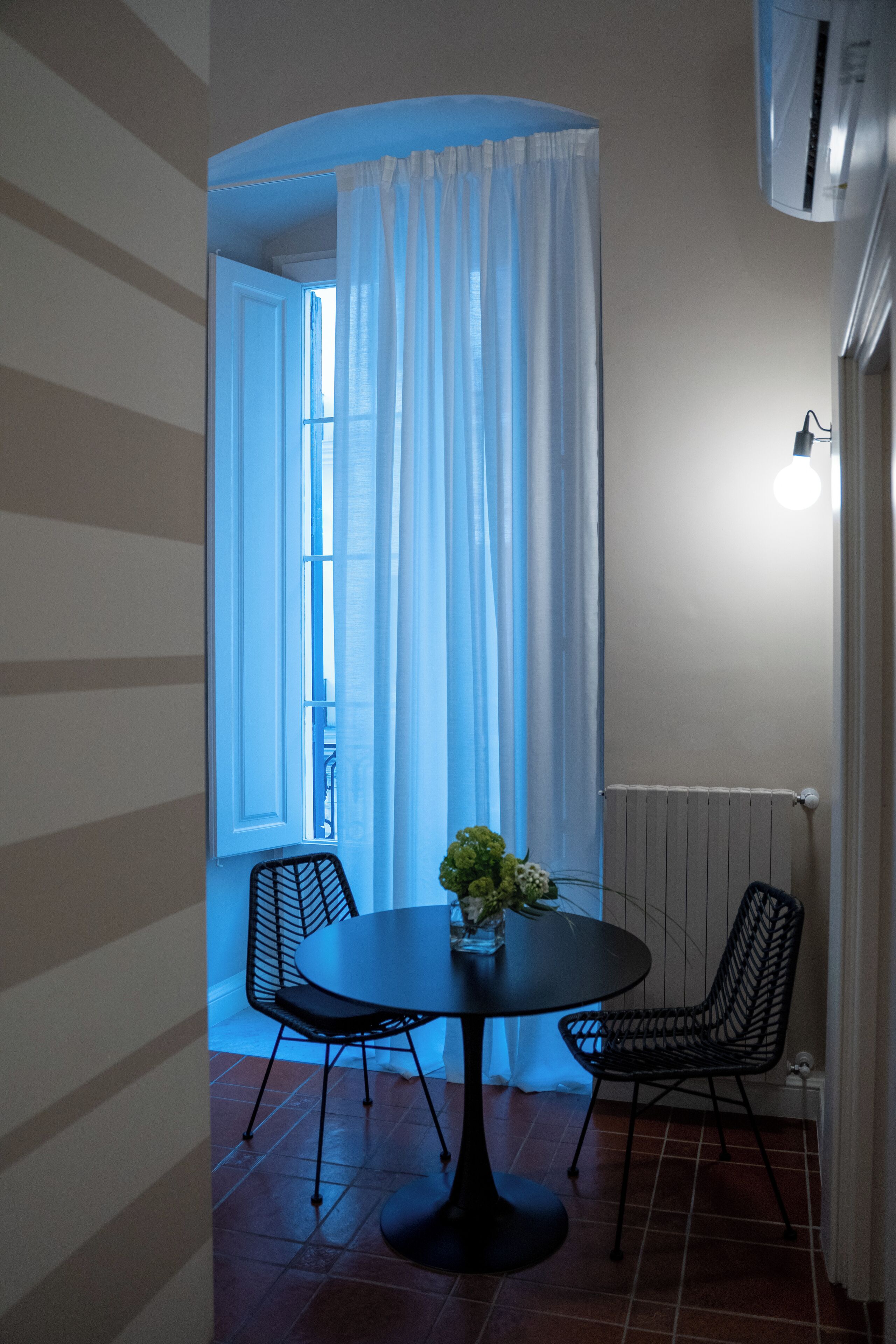 A dining area with a round table, two chairs, a vase with flowers, and a window with curtains.