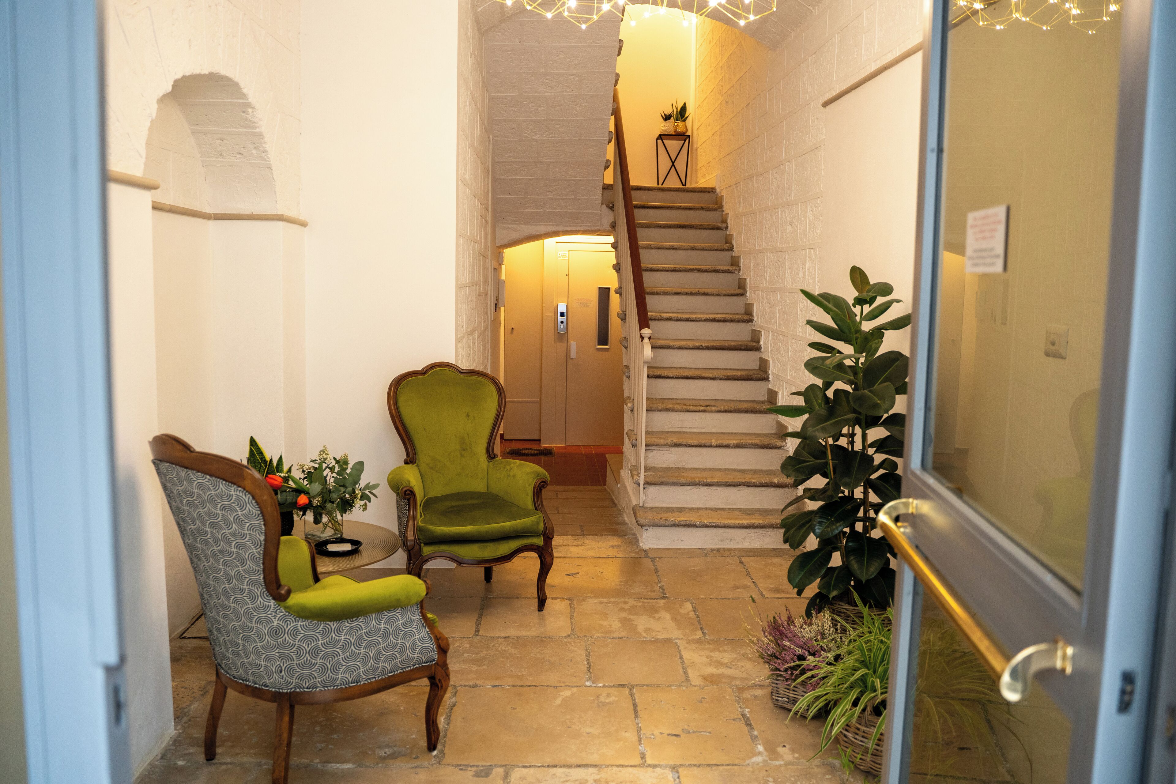 A narrow hallway with a staircase leading to an upper floor. There are two chairs with cushions, a potted plant, and a small table with a vase. The walls are white, and the floor is made of stone tiles.