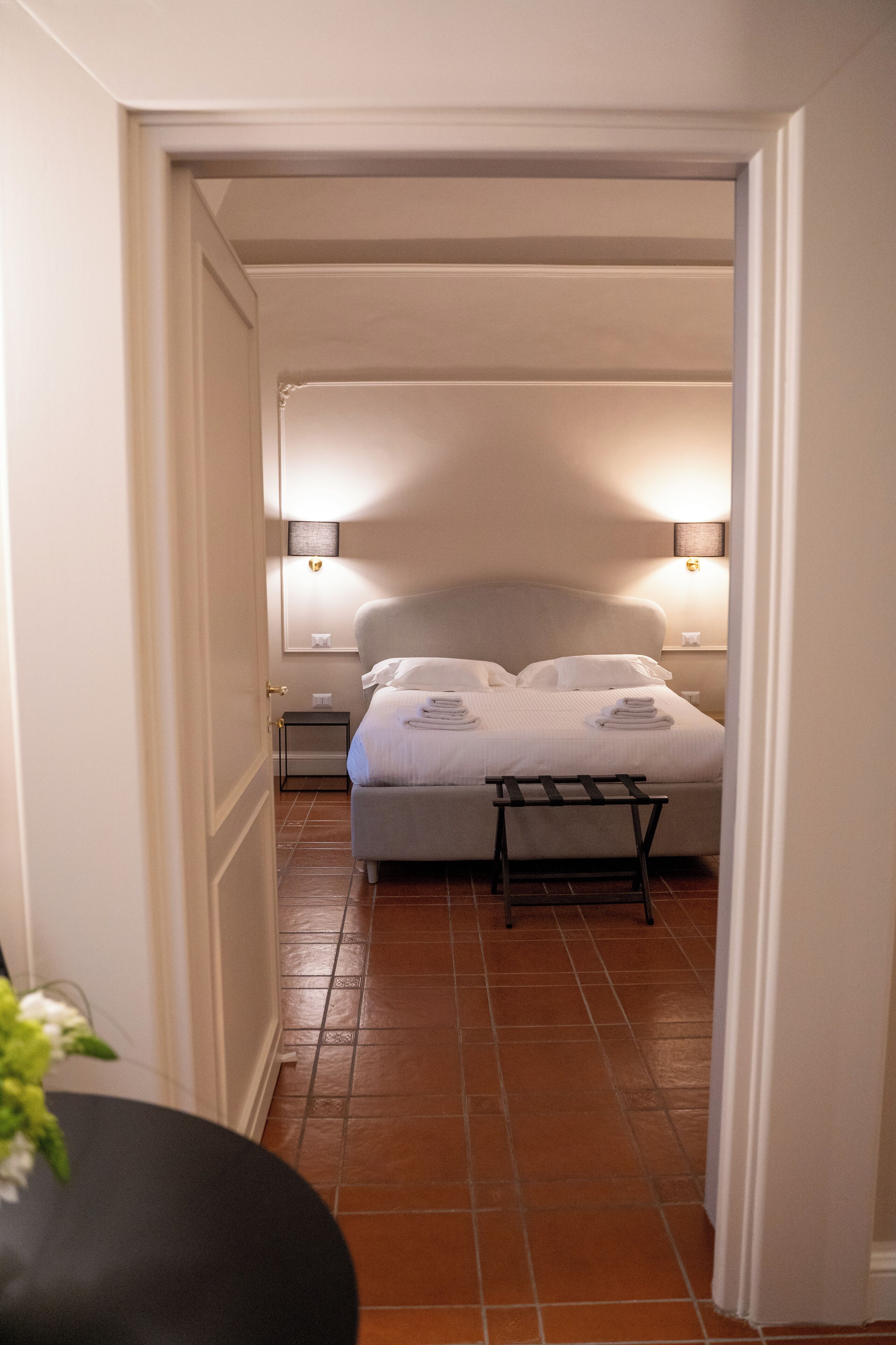 A hotel room with a bed, bedside lamps, a small table, and a tiled floor.