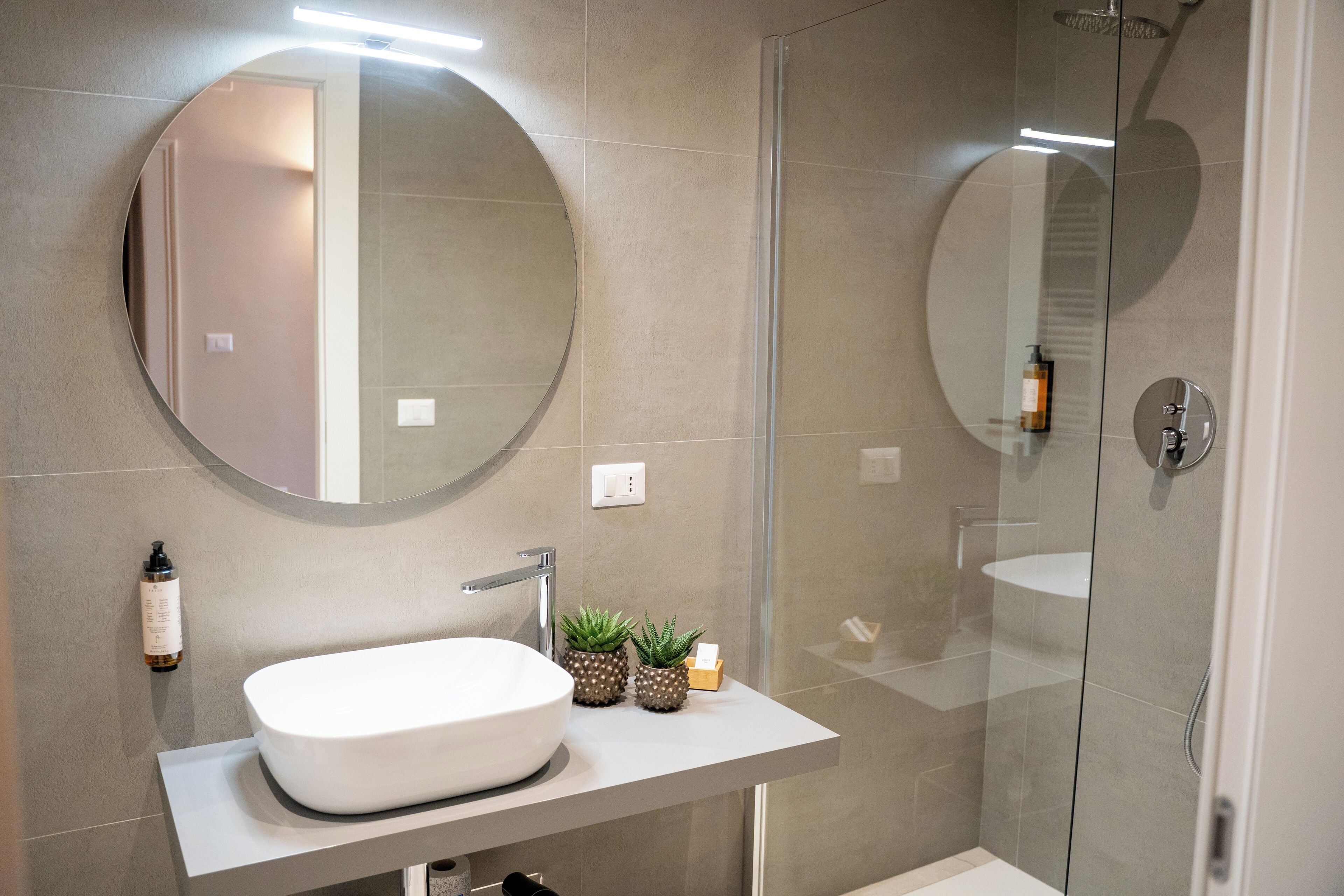 A modern bathroom with a round mirror, a white sink, and a glass-enclosed shower.