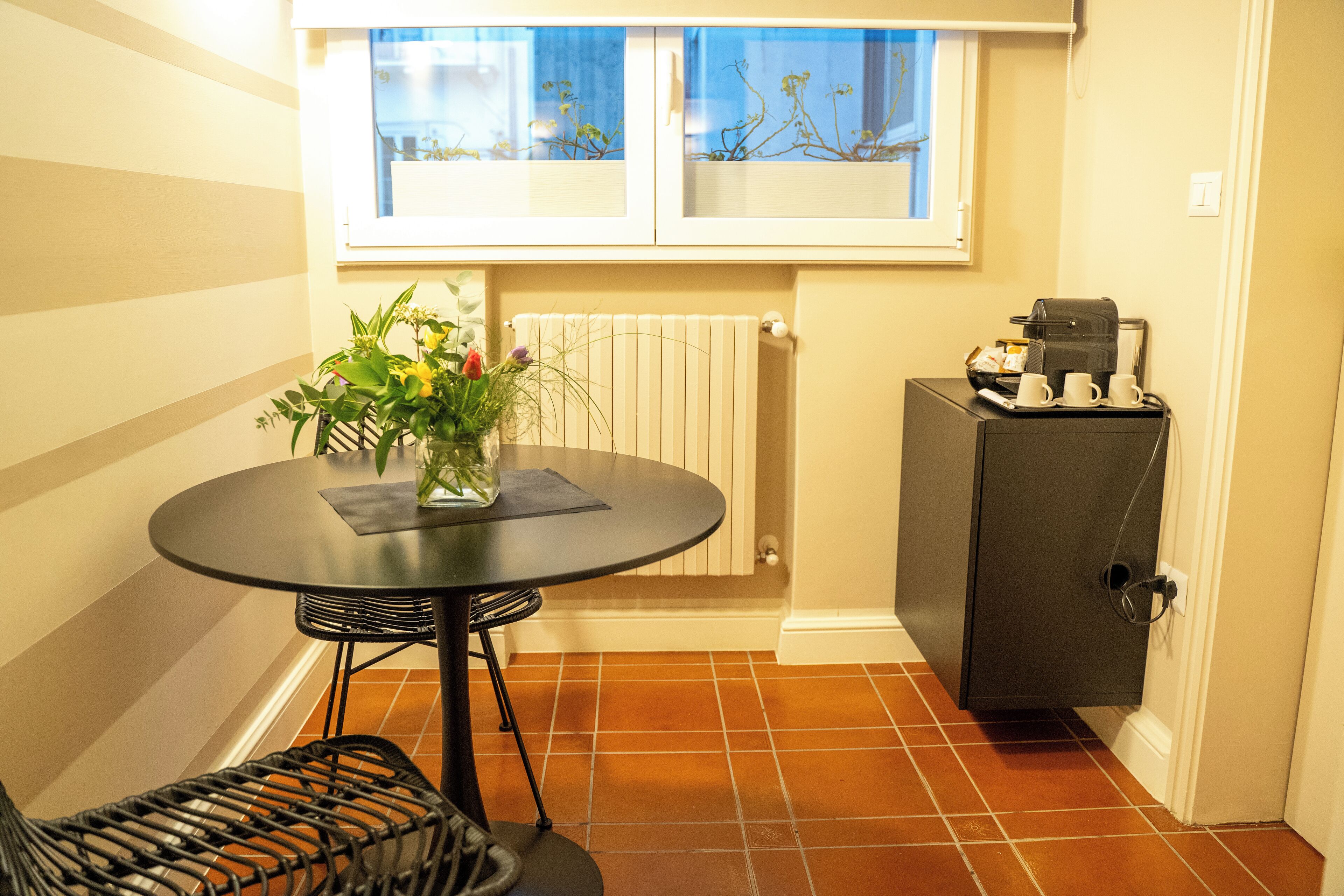 A small dining area with a round table, a chair, a potted plant, a radiator, and a coffee machine.