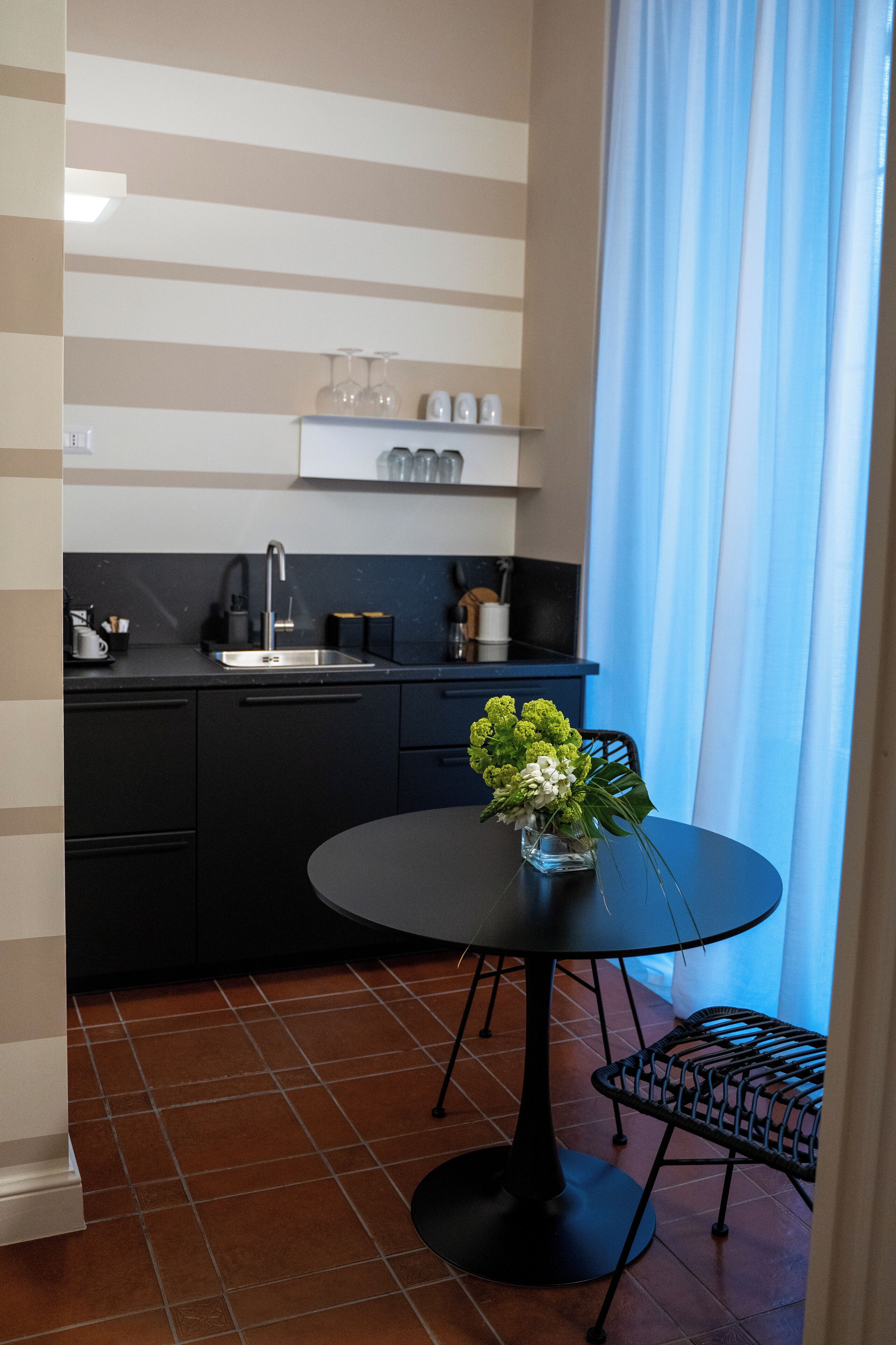 Una cocina compacta con encimera negra, una mesa redonda con un jarrón de flores y una ventana con cortinas azules.