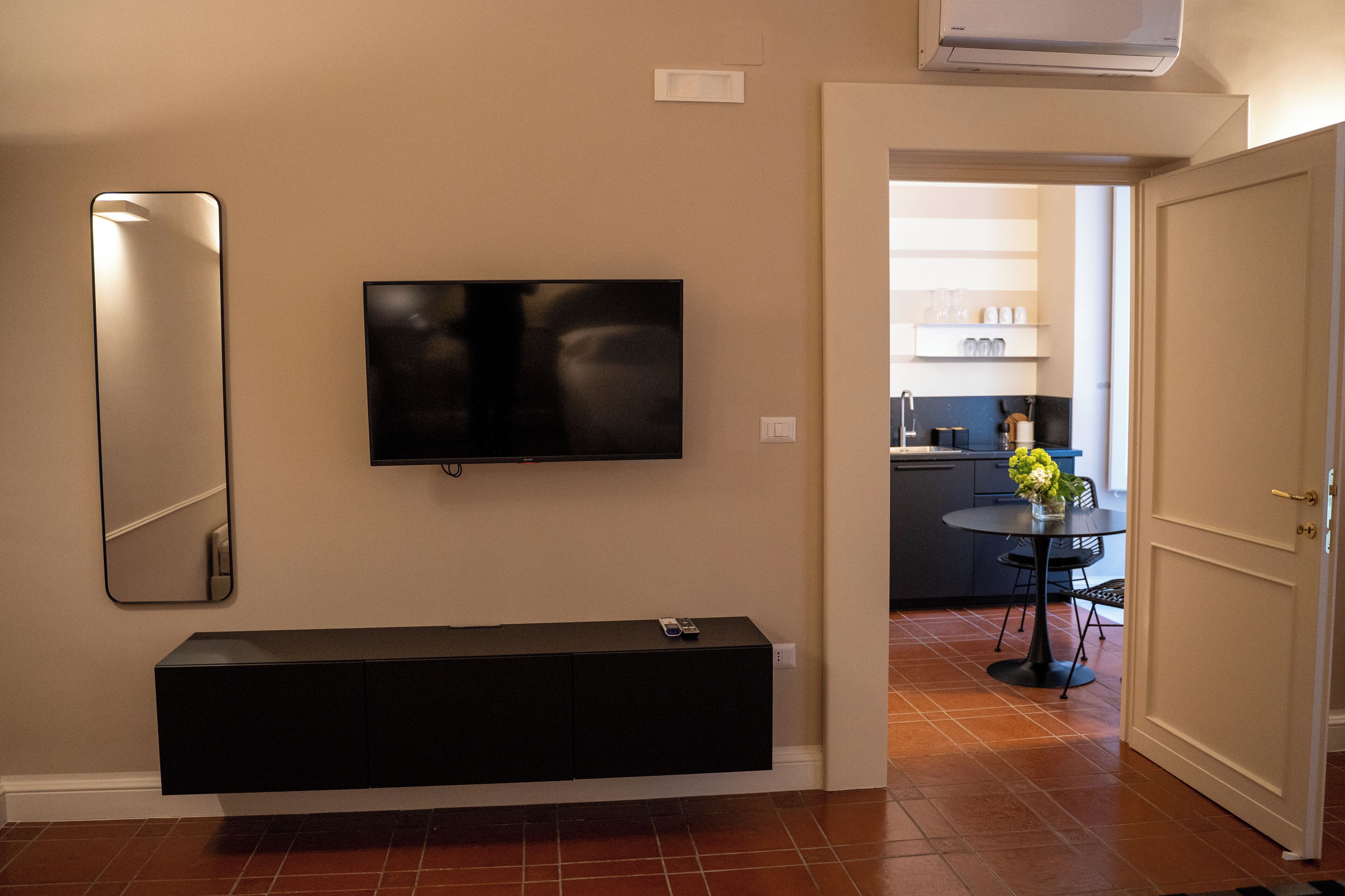A modern living room with a flat-screen TV mounted on the wall, a black entertainment unit below, and a kitchen visible through an open door.