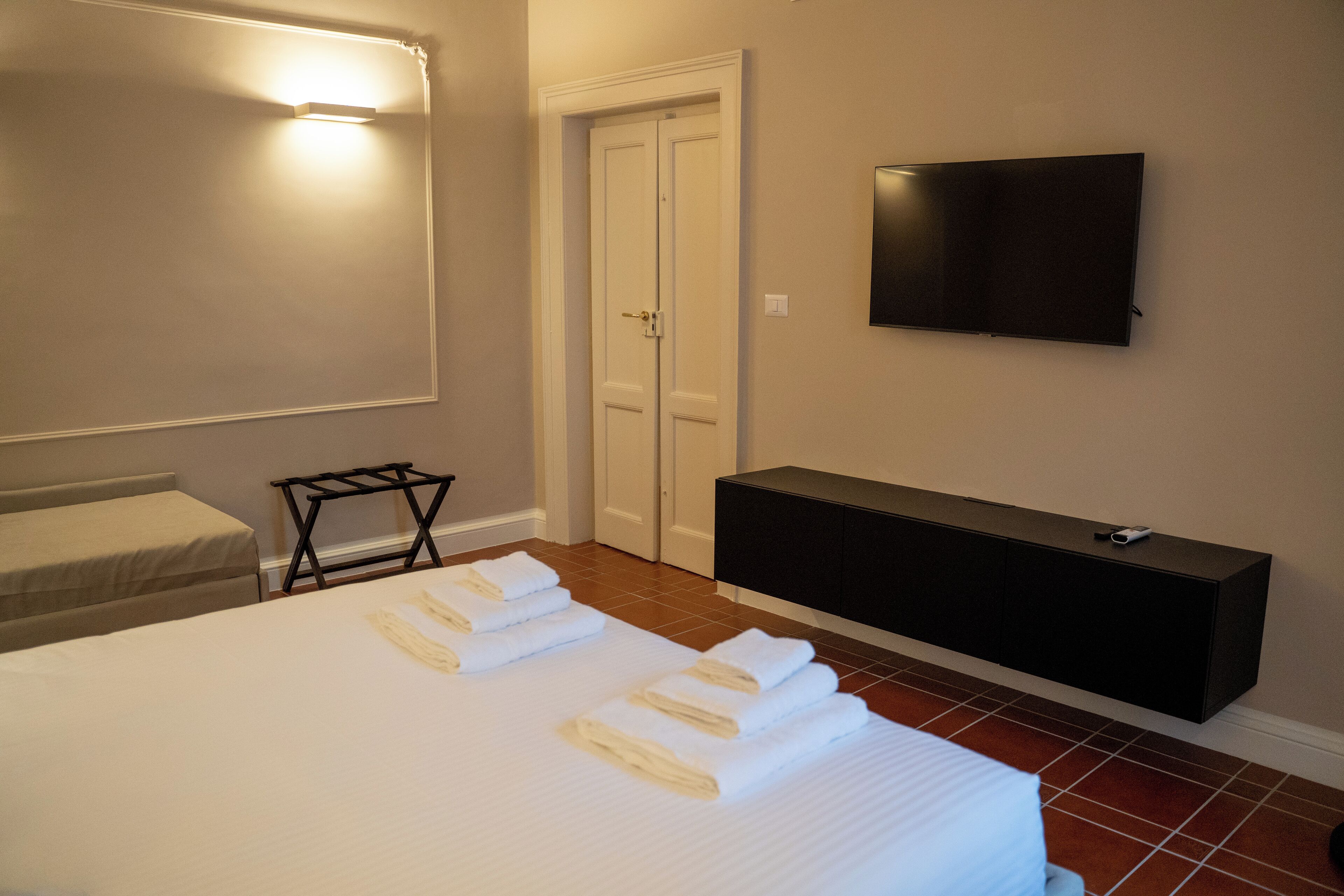 Una habitación de hotel con una cama, un televisor montado en la pared, una mesita consola negra y una puerta con interruptor de luz.