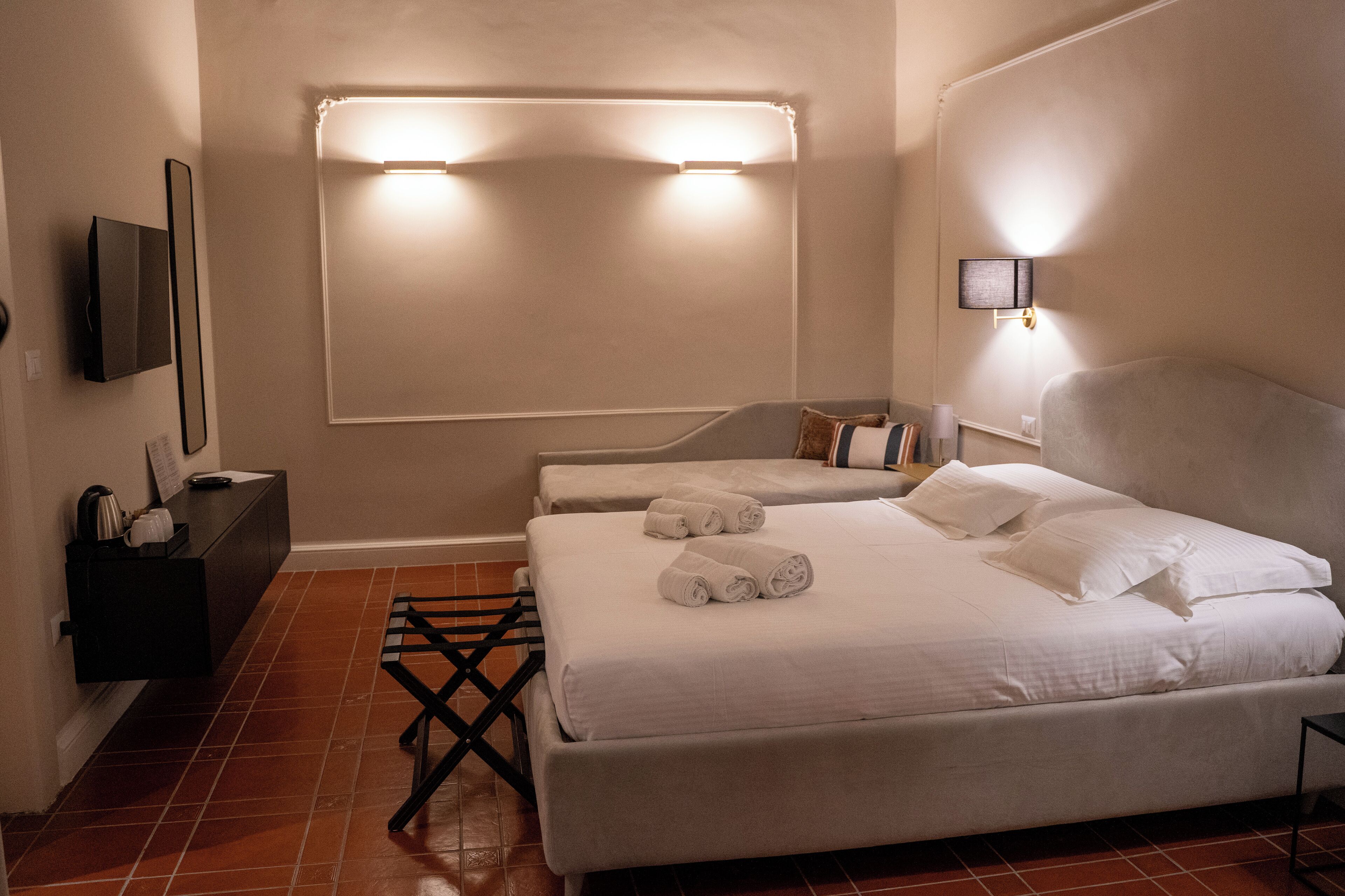 A hotel room with two beds, a flat-screen TV mounted on the wall, a small table with a kettle, and a tiled floor.