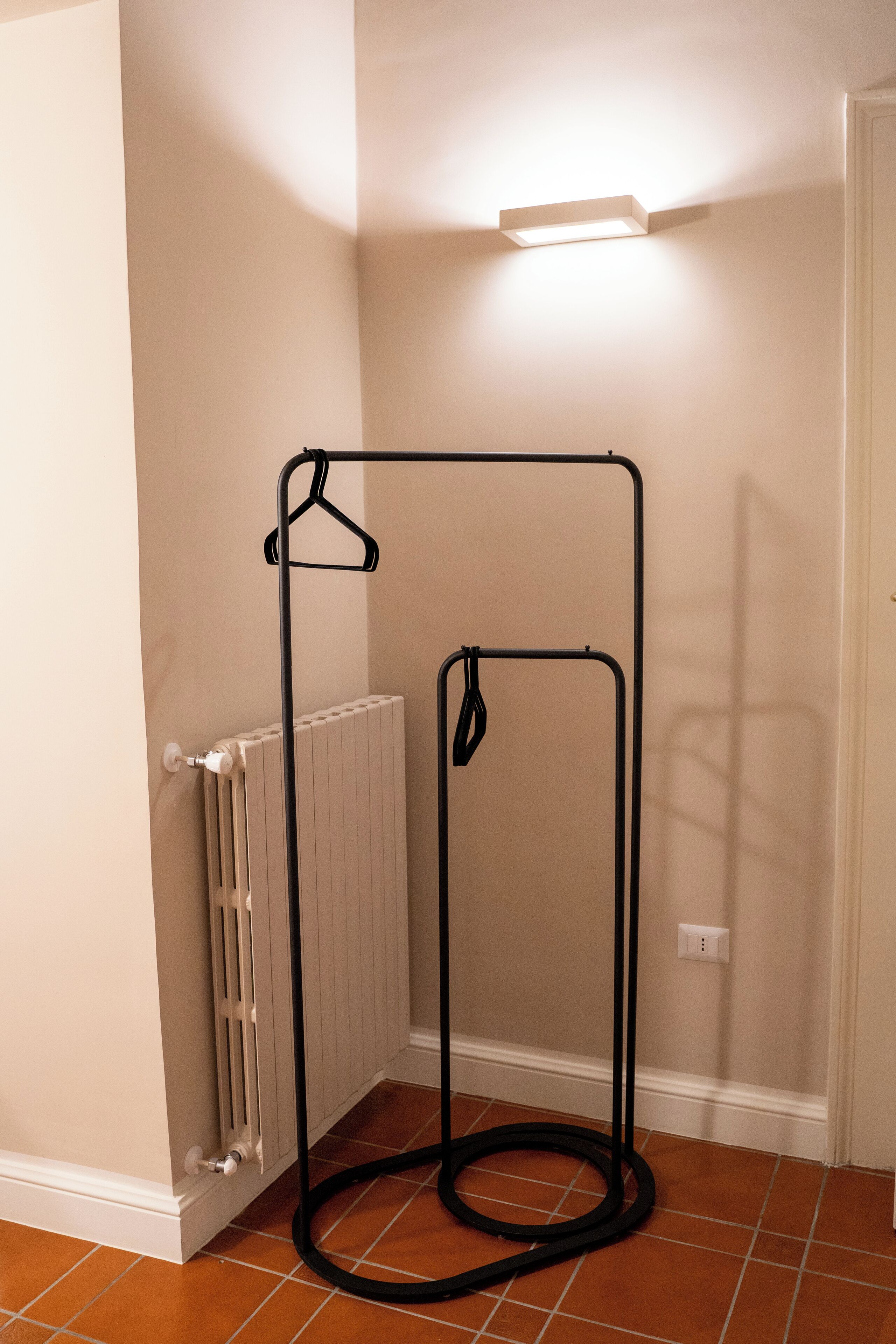 A corner room with a radiator, a black metal hanger rack, and a light fixture on the wall.