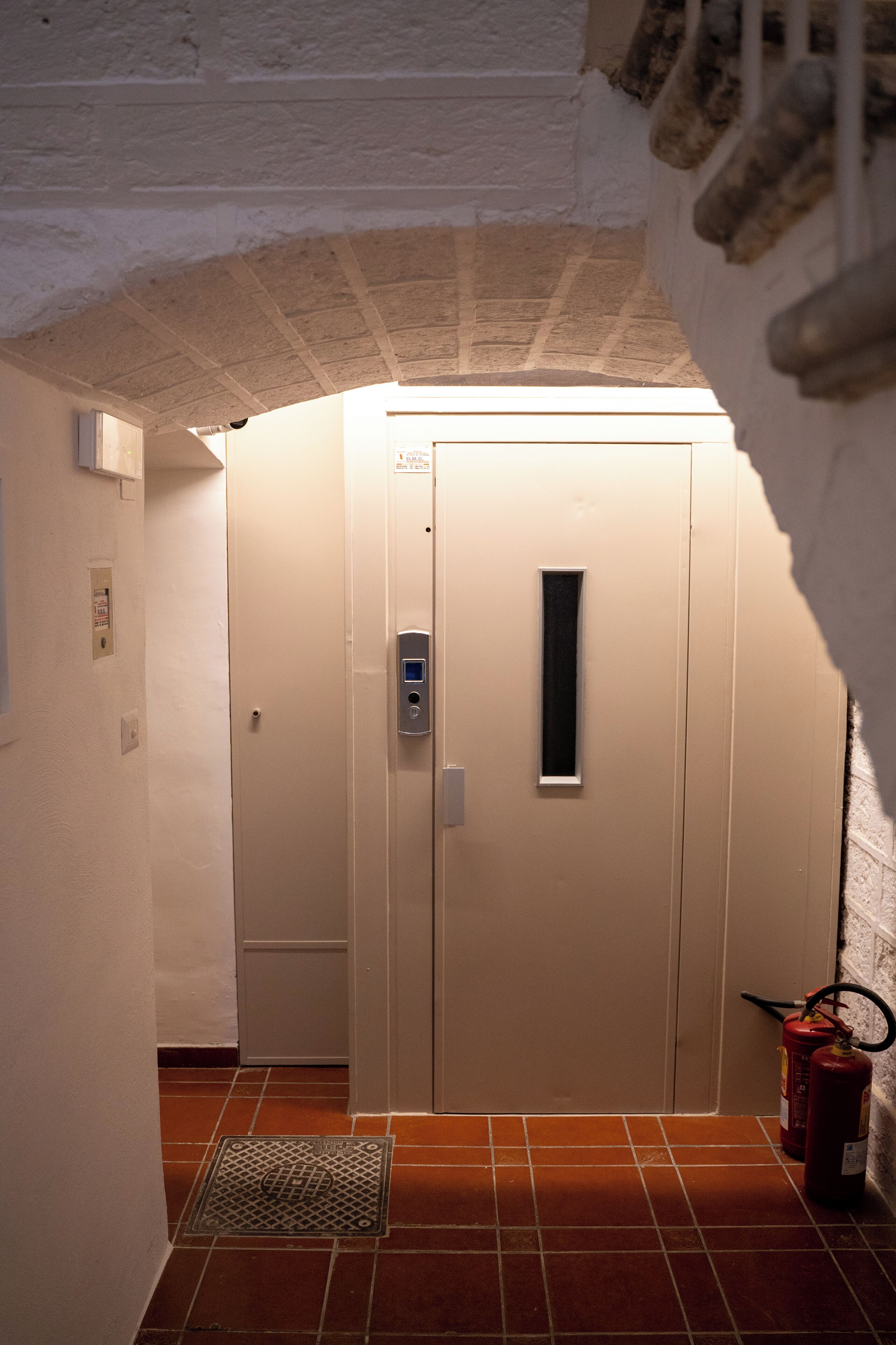 A narrow hallway with a tiled floor, a door with a handle, a fire extinguisher, and a small vent.