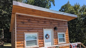 Une cabane en rondins avec une véranda, deux fenêtres, une porte ornée d’une guirlande et un drapeau américain.