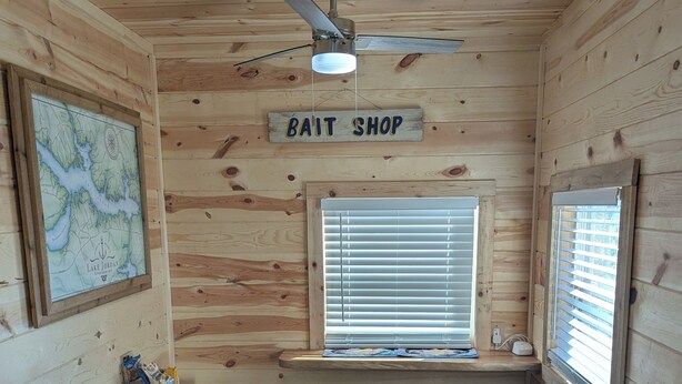 L’intérieur d’un chalet en bois, avec un ventilateur de plafond, une carte accrochée au mur, une enseigne indiquant un magasin de vente d’appâts, deux tabourets, une fenêtre avec des stores et un petit bureau avec des papiers.