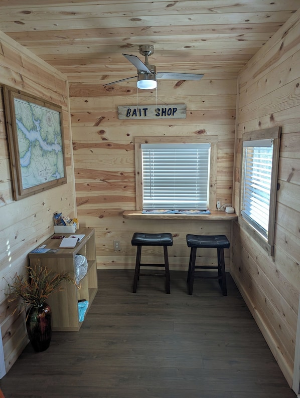 L’intérieur d’un chalet en bois, avec un ventilateur de plafond, une carte accrochée au mur, une enseigne indiquant un magasin de vente d’appâts, deux tabourets, une fenêtre avec des stores et un petit bureau avec des papiers.