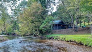 Eine Hütte am Flussufer mit einer Terrasse, umgeben von dichtem Wald und einem klaren Bach.
