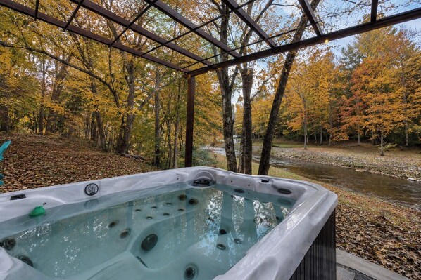 A hot tub in a covered outdoor area with a view of a forest and a stream.