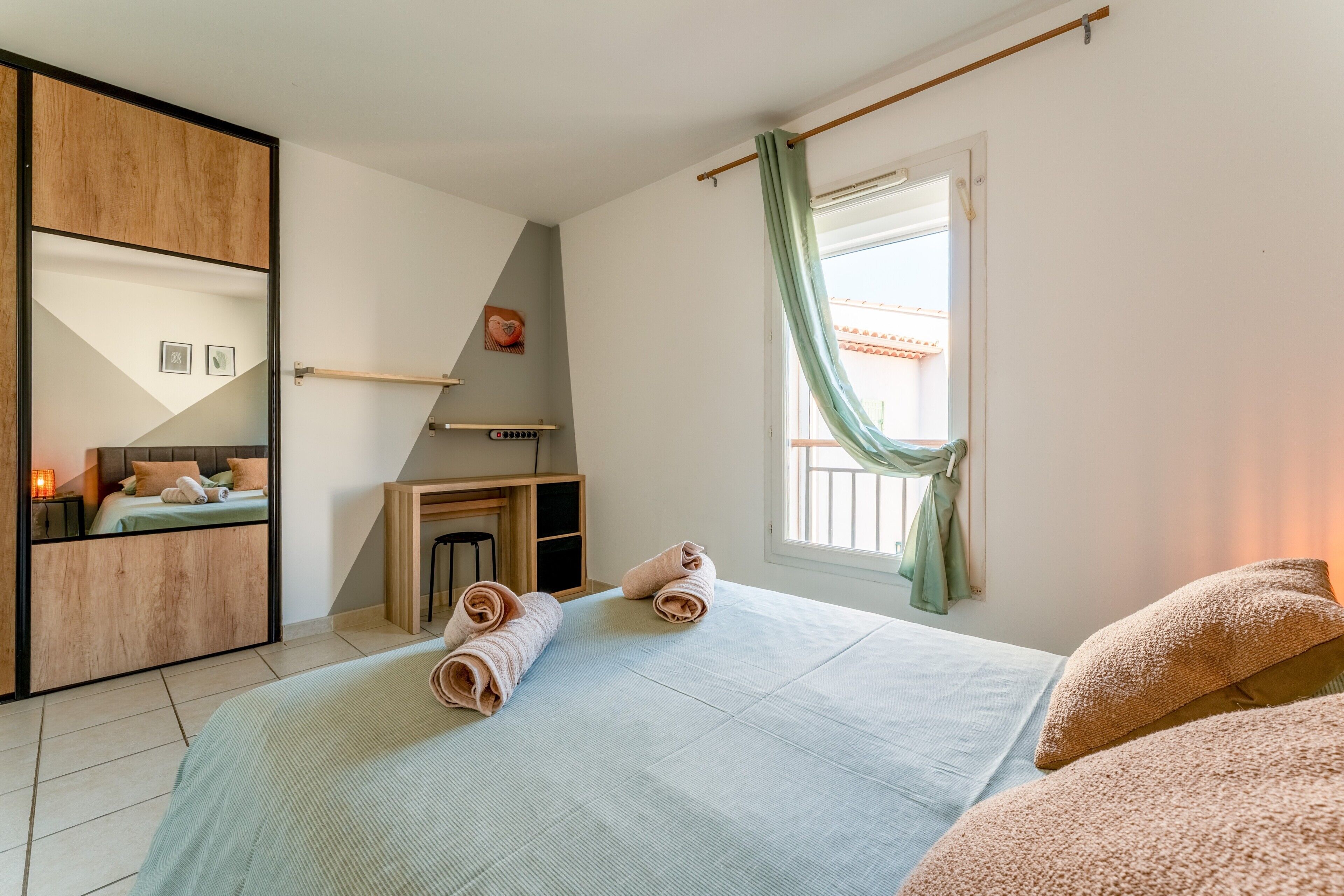 Une chambre à coucher avec un lit, un bureau, une chaise, une fenêtre avec des rideaux et un miroir.