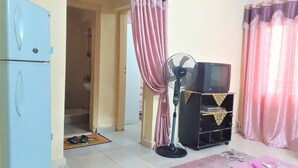 A living room with a blue refrigerator, a standing fan, a television on a wooden stand, and a pink curtain. - Furnished apartment in Minya with archaeological tours (Minya Governorate)