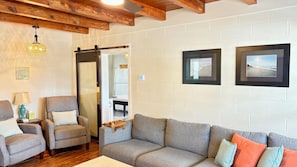 Un salon avec un canapé gris, deux fauteuils, un plafond en bois et deux tableaux encadrés au mur.