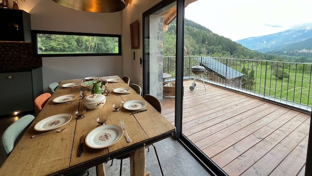 A dining area with a wooden table set for four, a large window, and a view of a mountainous landscape. - Chalet at the foot of the ski resorts with spa (Landry)