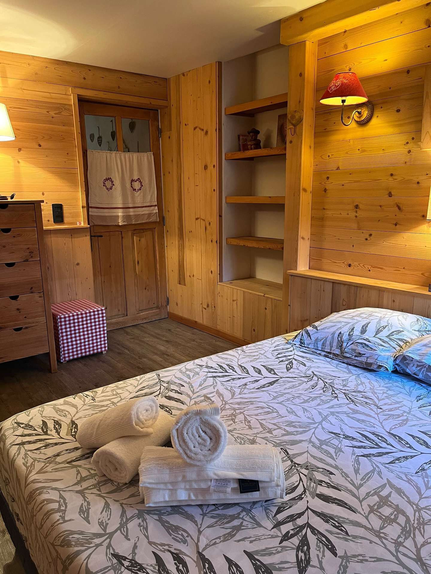 A cozy room with a bed, a wooden wardrobe, a small table, and a shelf unit.