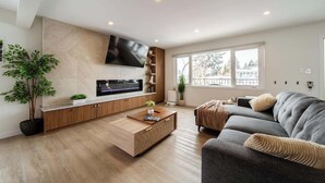 Una sala de estar moderna con un televisor de pantalla plana montado en la pared, un sofá gris, una mesa de centro de madera y un ventanal con vistas al exterior.