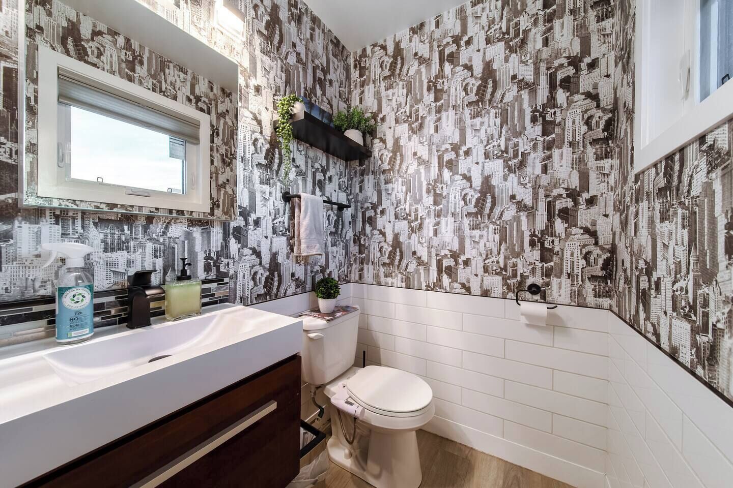 Een badkamer met stadsgezicht behang, een witte wastafel, een toilet en een raam met uitzicht.