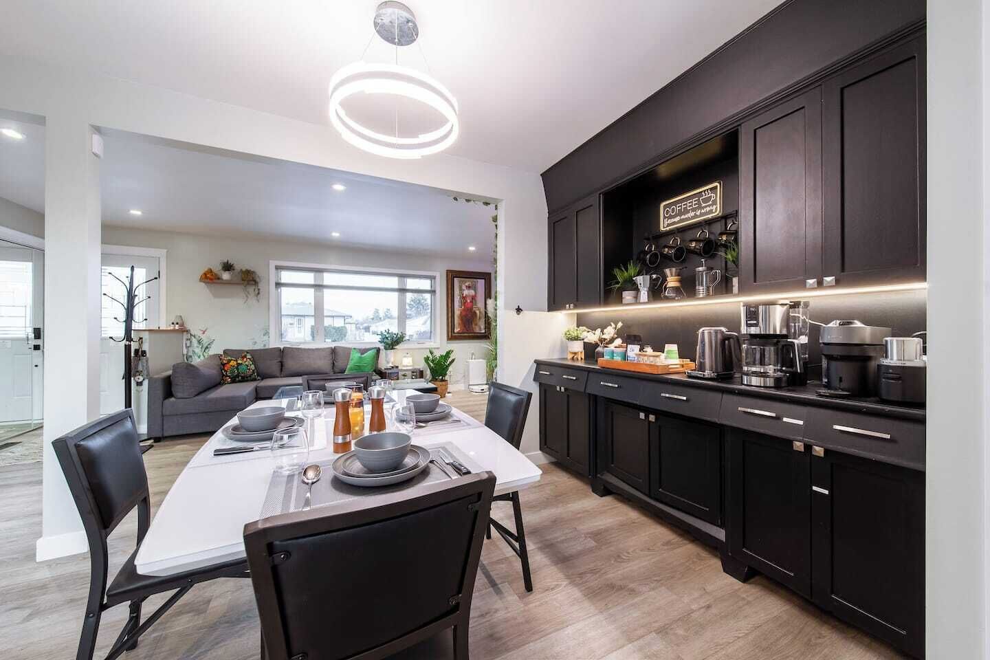 A modern kitchen with dark cabinetry, a dining area with a white table set for four, and a living room with a gray sofa and green cushions.