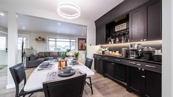 A modern kitchen with dark cabinetry, a dining area with a white table set for four, and a living room with a gray sofa and green cushions.
