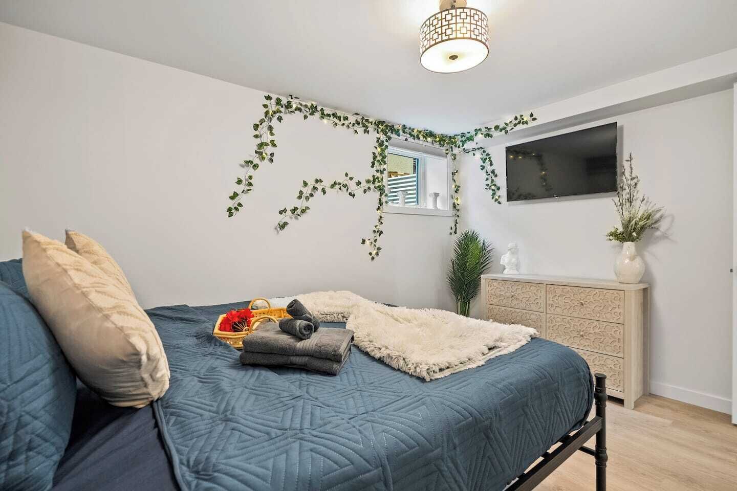 A bedroom with a bed, pillows, a blanket, a dresser, a TV, and a plant.