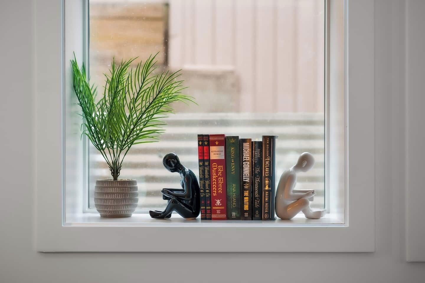 Een vensterbank met een bloempot, een zwart beeldhouwwerk en een rij boeken.