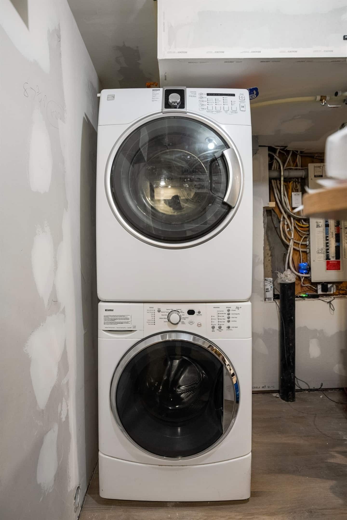 A stacked washer and dryer unit in a room with exposed electrical wiring and unfinished walls.