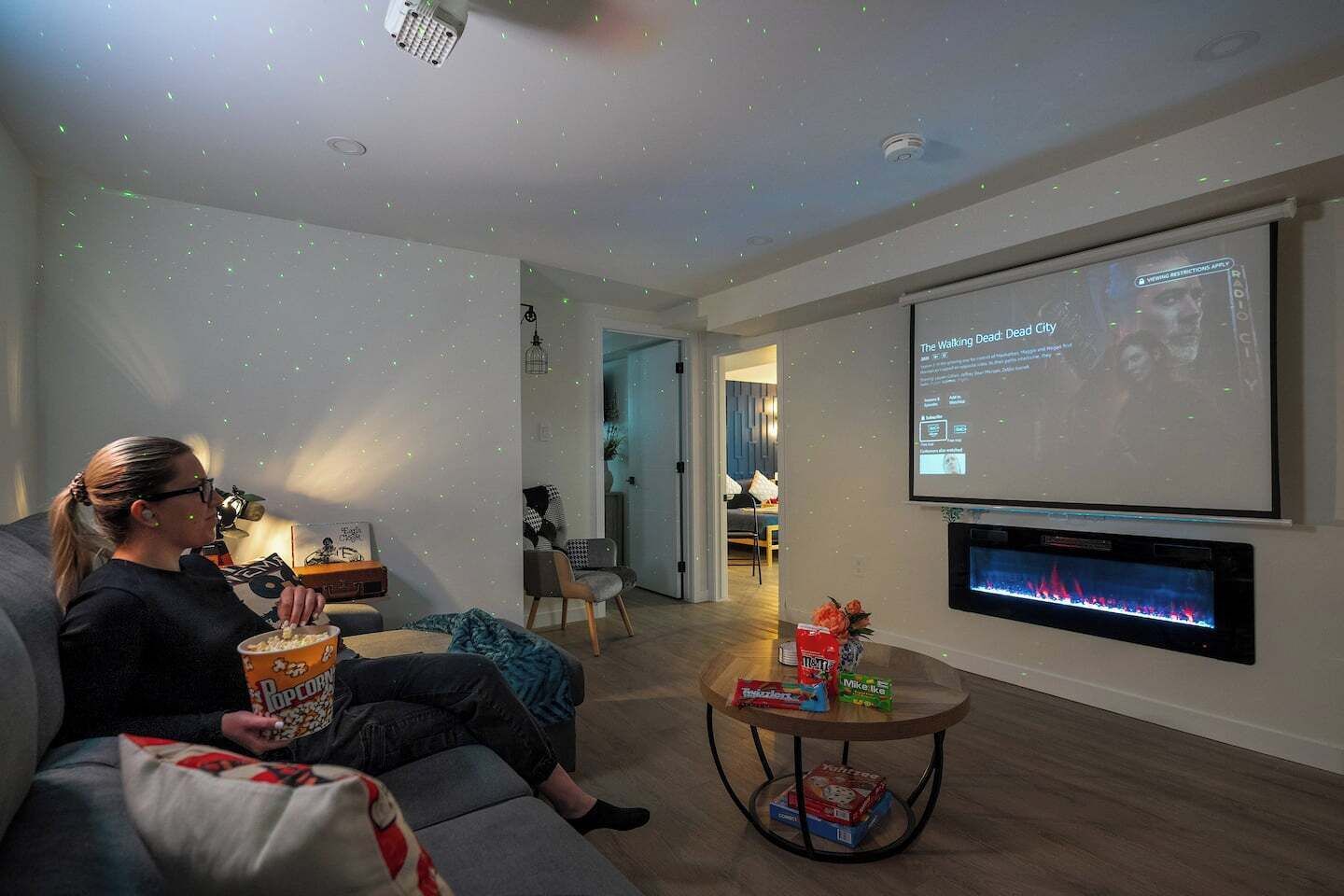 A person sitting on a couch with a popcorn bucket, a TV screen displaying a movie, and a projector screen showing a movie title.