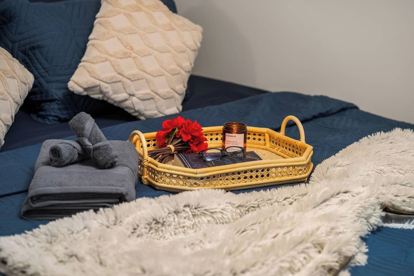 A neatly made bed with a blue comforter, a white fluffy throw, and a wicker tray with a candle, flowers, and a book.