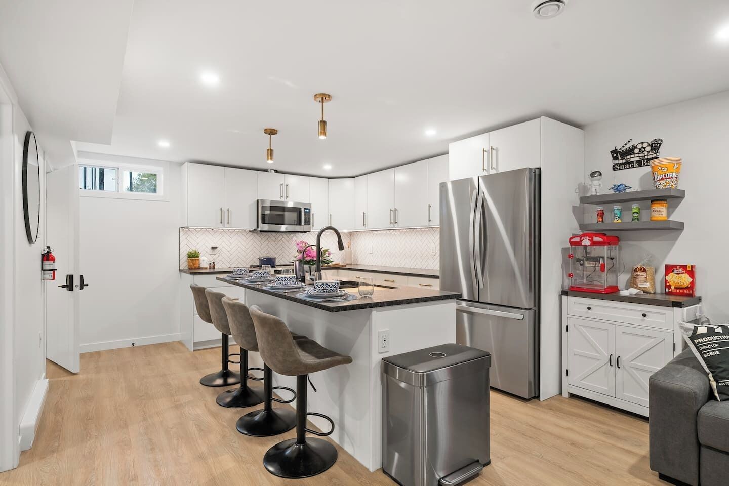 A modern kitchen with white cabinets, a stainless steel refrigerator, a microwave, and a coffee machine. There are three bar stools and a trash bin.