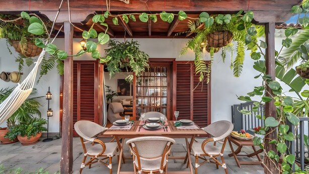 Een eetgedeelte met een houten tafel voor vier personen, omringd door rieten stoelen, onder een houten pergola met hangende planten.