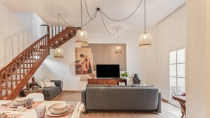 Een ruime woonkamer met een houten trap, een grijze bank, een flatscreen televisie en hanglampen.