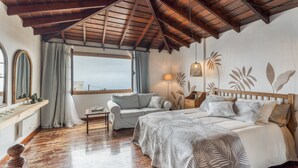 Een slaapkamer met een houten plafond, een bed met een sprei met bladmotief, een bank, een kleine tafel en een raam met gordijnen.