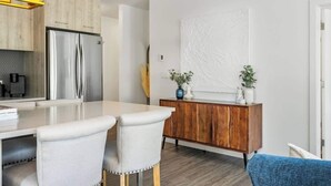 A modern kitchen with a stainless steel refrigerator, wooden cabinets, and a white countertop. There is a dining area with two chairs and a wooden sideboard with decorative items. - Walk to cafes, Safeway, and trails from this stylish Whyte Ave groundfloor suite (Edmonton)