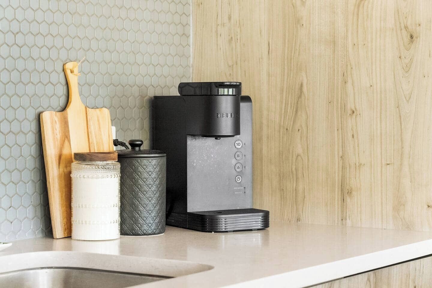 A kitchen counter with a coffee maker, a wooden cutting board, a white ceramic jar, and a textured metal container.