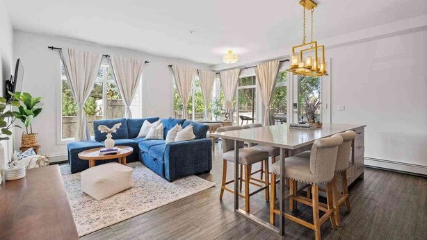 A modern living room with a blue sectional sofa, a dining table with bar stools, and large windows with curtains. - Walk to cafes, Safeway, and trails from this stylish Whyte Ave groundfloor suite (Edmonton)