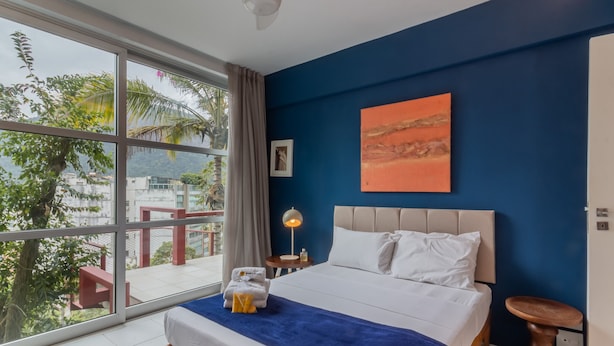 Una habitación de hotel con un ventanal grande, una cama con cubrecama azul, una mesita de noche y un cuadro en la pared.