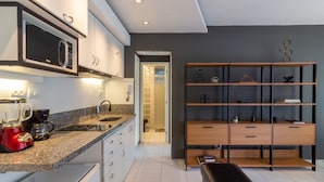 Una cocina moderna con microondas integrado, cafetera y licuadora. Hay una estantería de madera con objetos decorativos y un banco.