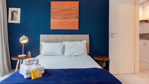 Una habitación de hotel con una cama, una mesita de noche con lámpara, un cuadro en la pared y una puerta que da al baño.