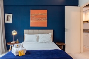 Una habitación de hotel con una cama, una mesita de noche con lámpara, un cuadro en la pared y una puerta que da al baño.