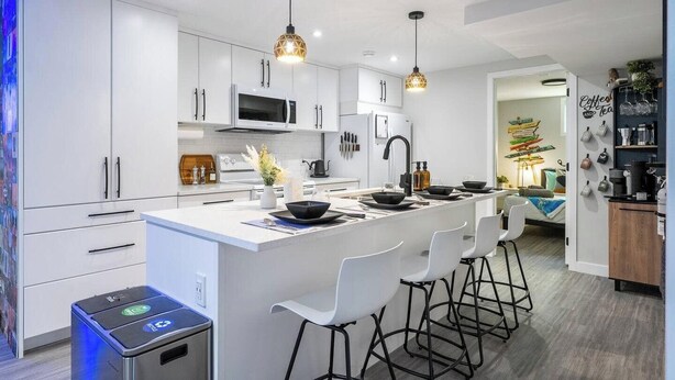 Een moderne keuken met witte kasten, een centraal keukenblok met barkrukken, hanglampen en een koffiehoek.