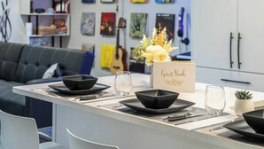 Een moderne eetruimte met een minimalistische tafel gedekt voor vier personen, met zwarte kommen en glazen. Er ligt een gastenboek op de tafel, en op de achtergrond staat een bank en een muur versierd met verschillende ingelijste kunstwerken.