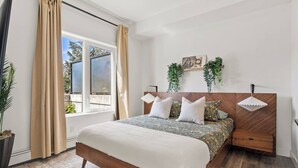A bedroom with a large bed, a wooden headboard, a bedside table, a window with curtains, and a hanging plant.