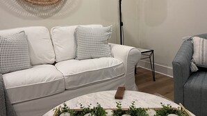 Una sala de estar con un sofá blanco, un sillón gris, una mesita redonda, una lámpara de pie y un tapiz tejido en la pared.