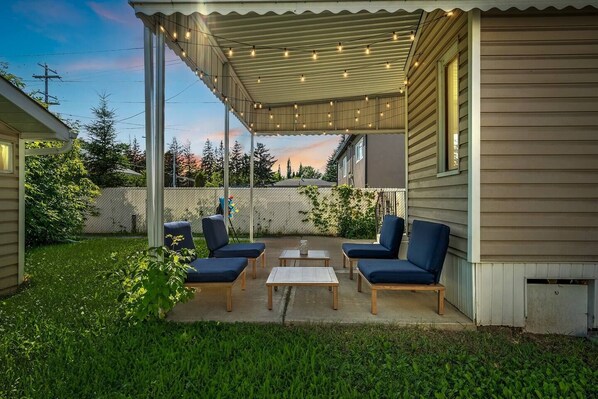 Eine überdachte Terrasse mit vier blauen Stühlen und einem kleinen Tisch, umgeben von einem Rasen und einem Holzzaun.