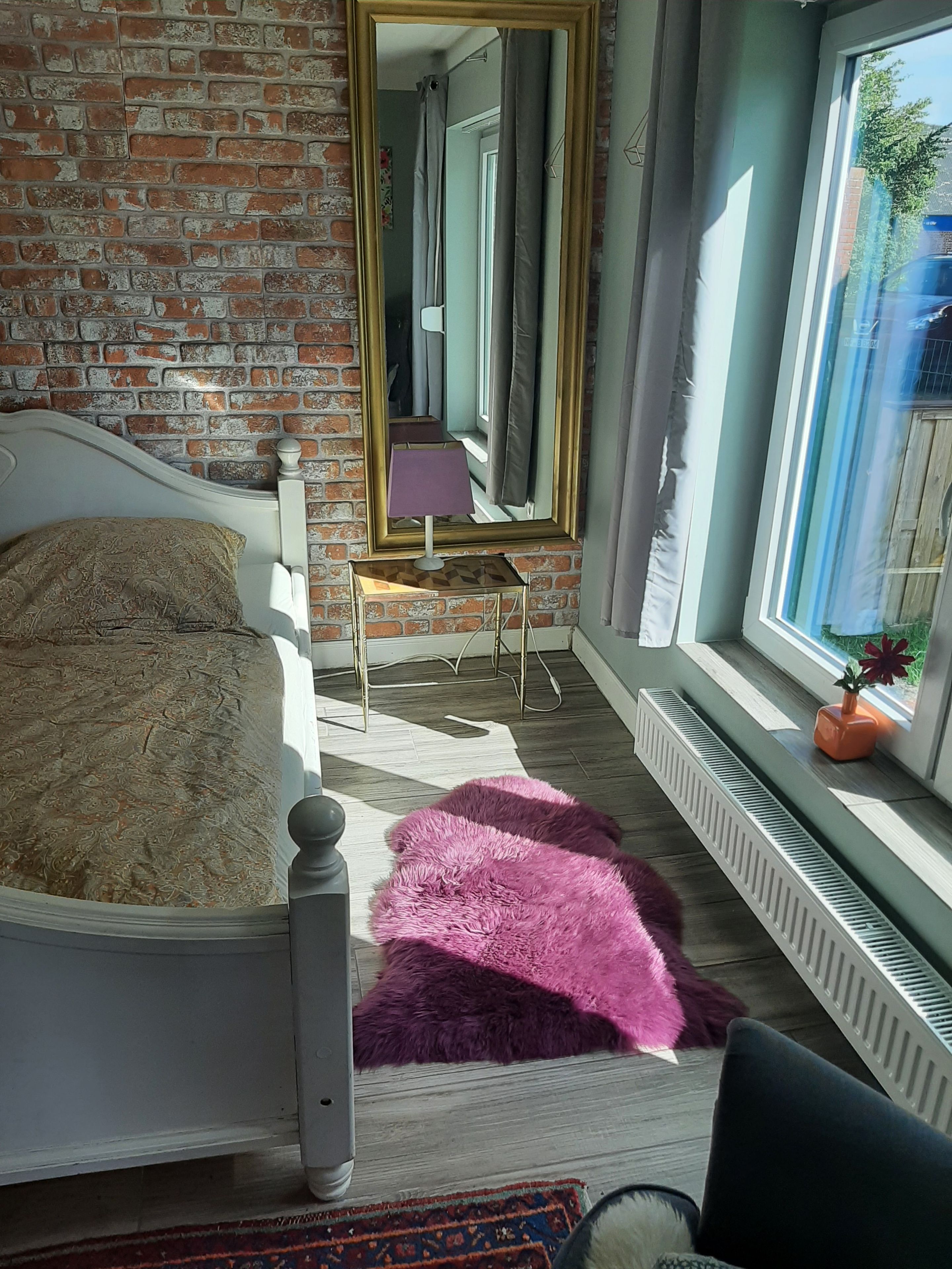 A bedroom with a brick wall, a bed with a patterned pillow, a small table with a lamp, a window with a view, and a radiator.